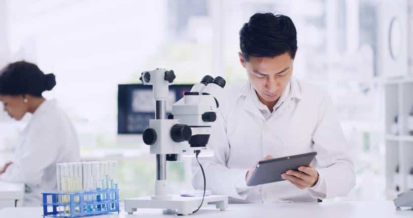 A researcher reviewing a tablet beside a microscope in a pharmaceutical laboratory