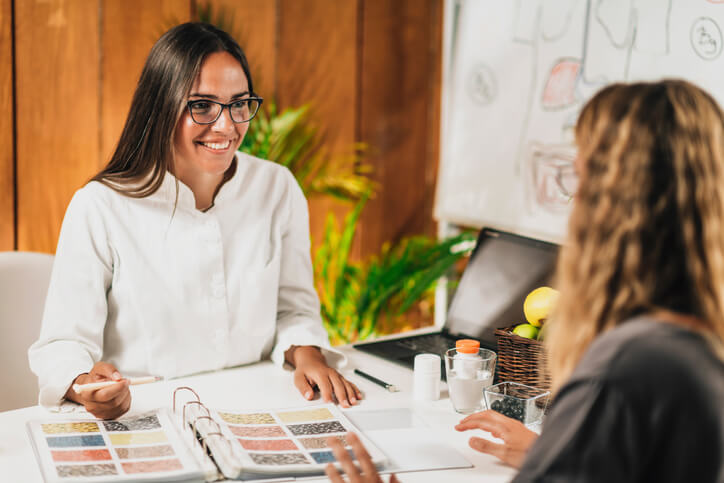 A smiling nutritionist wearing glasses explains food options to a client using a binder with seed and grain samples. The friendly consultation takes place in a bright office with healthy foods and a whiteboard behind.