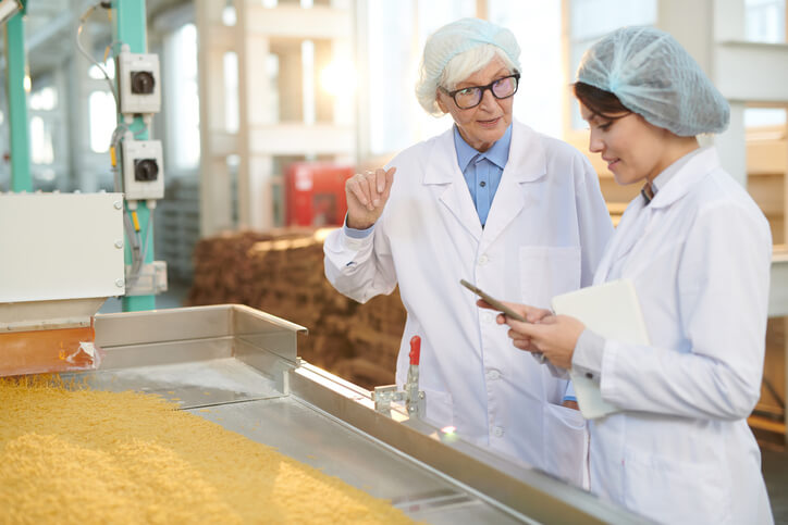 Two professionals in lab coats and hairnets discuss quality control in a food processing facility. One holds a mobile device and a notebook while the other gestures, overseeing pasta or grain production on a conveyor.